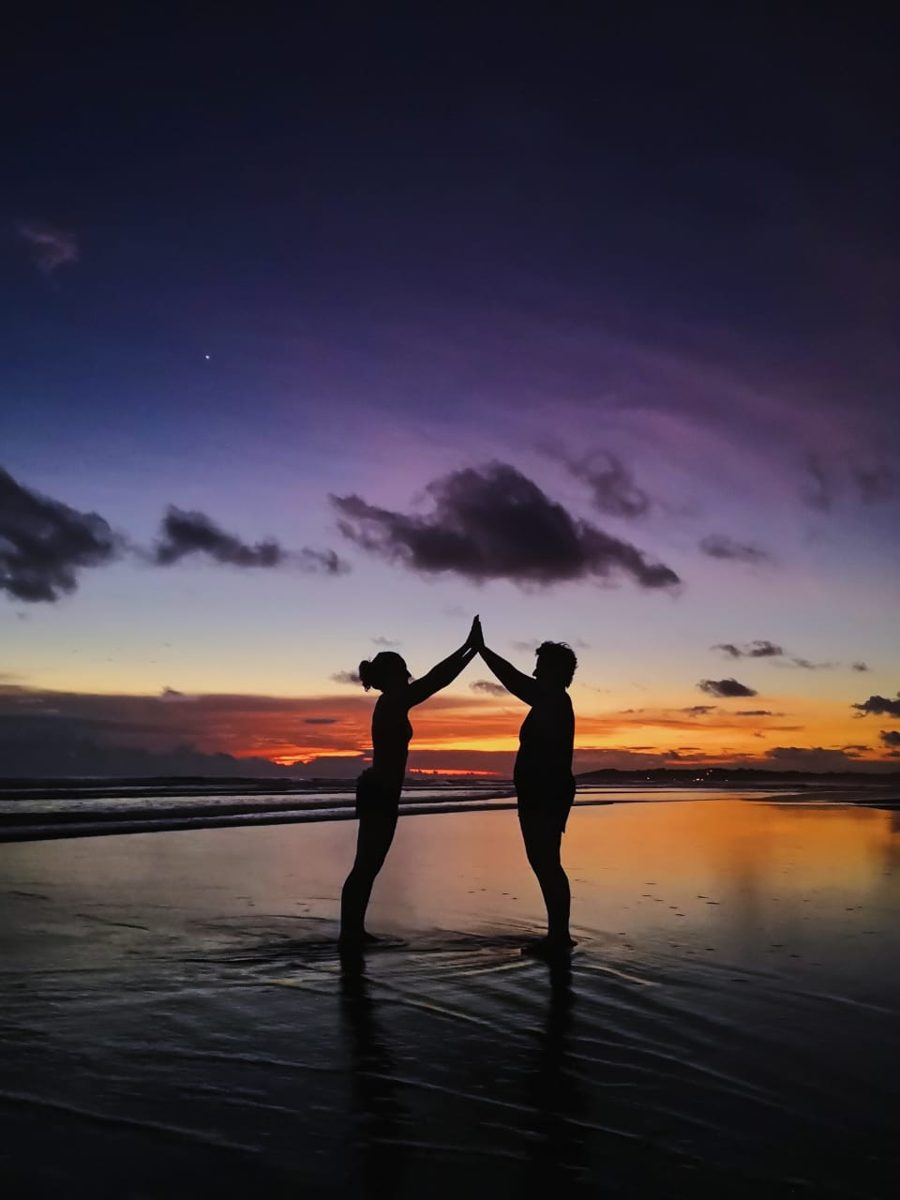 Dos mujeres tocándose las manos en alto frente al atardecer en la playa
